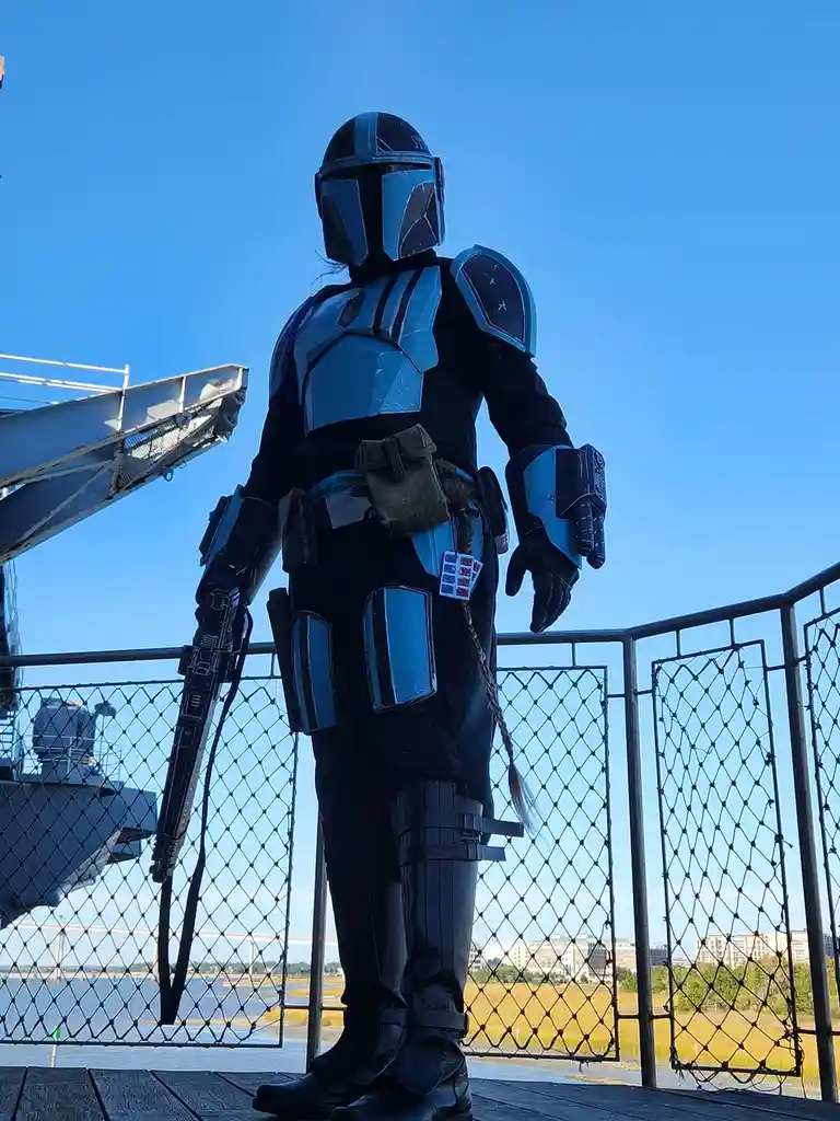 A person in a custom blue and black Mandalorian armor costume stands on a wooden deck with a chain-link fence, holding a…