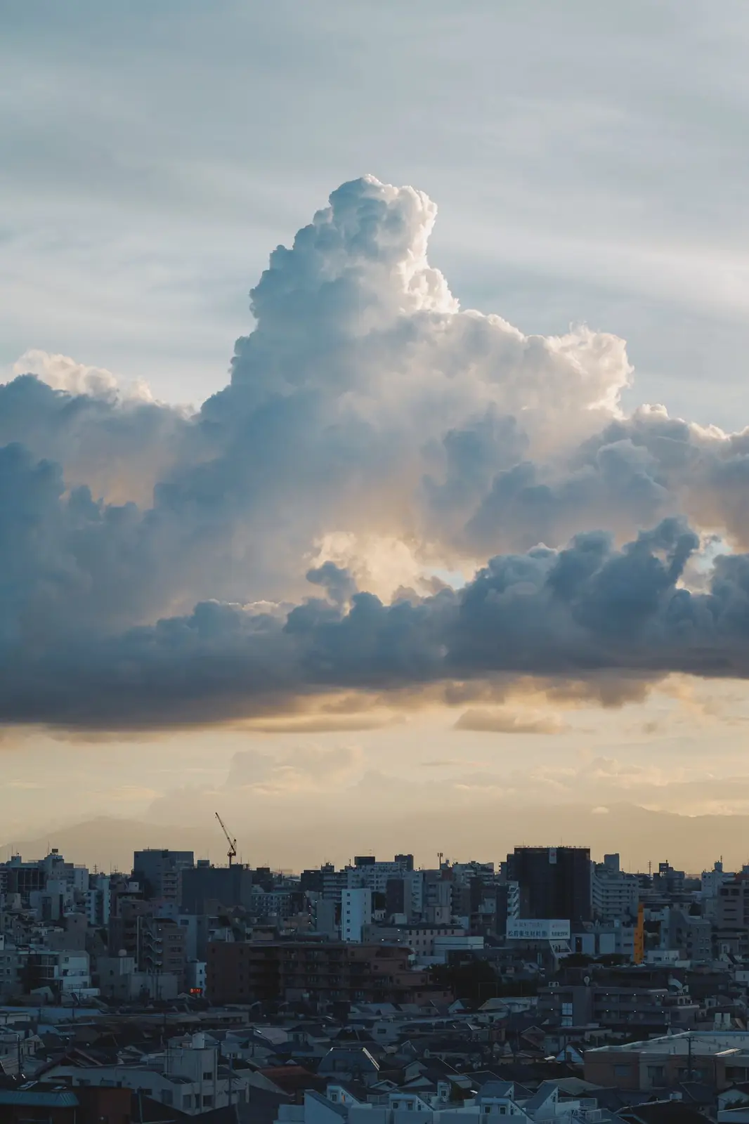 A dramatic cityscape photo shows a large, towering cloud formation over a dense urban area at sunset or sunrise, with…