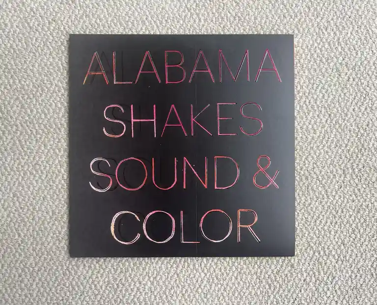 A square black album cover for Alabama Shakes' 'Sound & Color' lies on a light-colored textured carpet. The text is rendered…