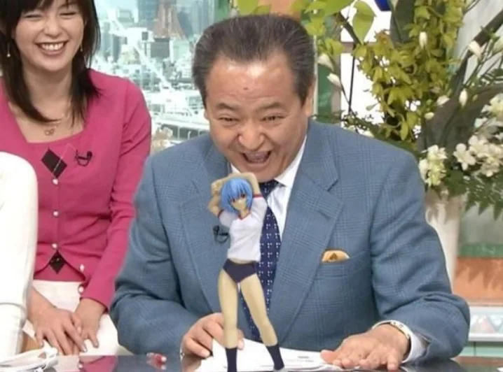 A man in a blue suit sits at a desk, laughing maniacally while holding up a small anime figurine of a girl with blue hair. A…