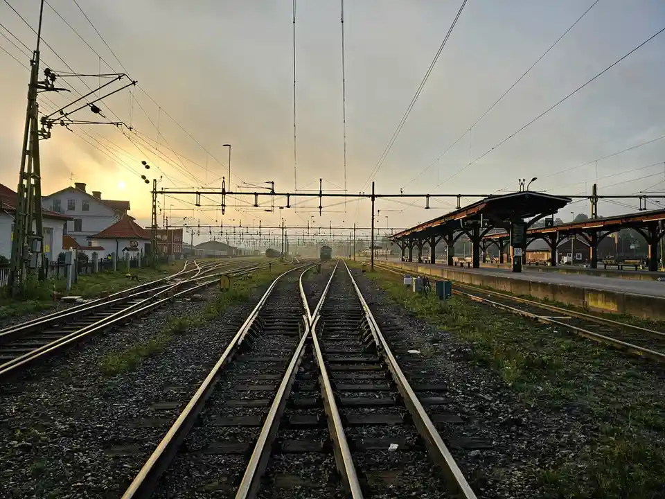 A photograph of a railway station at sunrise or sunset, with multiple tracks converging and diverging, overhead power lines…