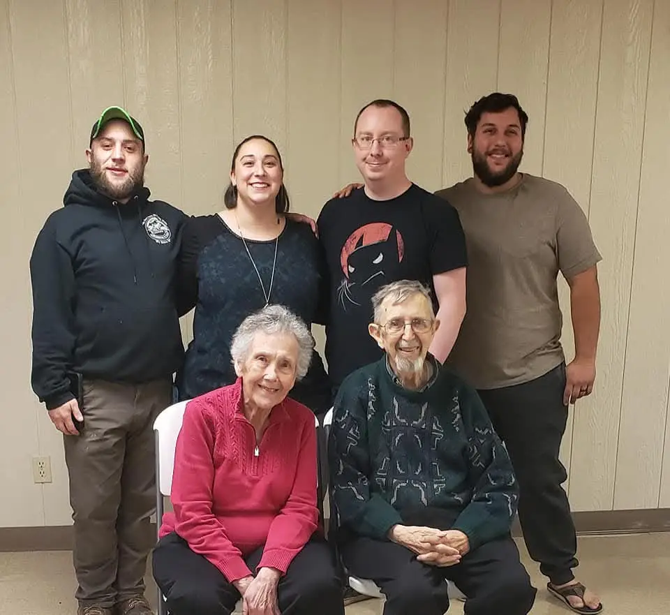 A group photo of six people, four younger adults standing behind two elderly people seated in front. The overlaid text reads…