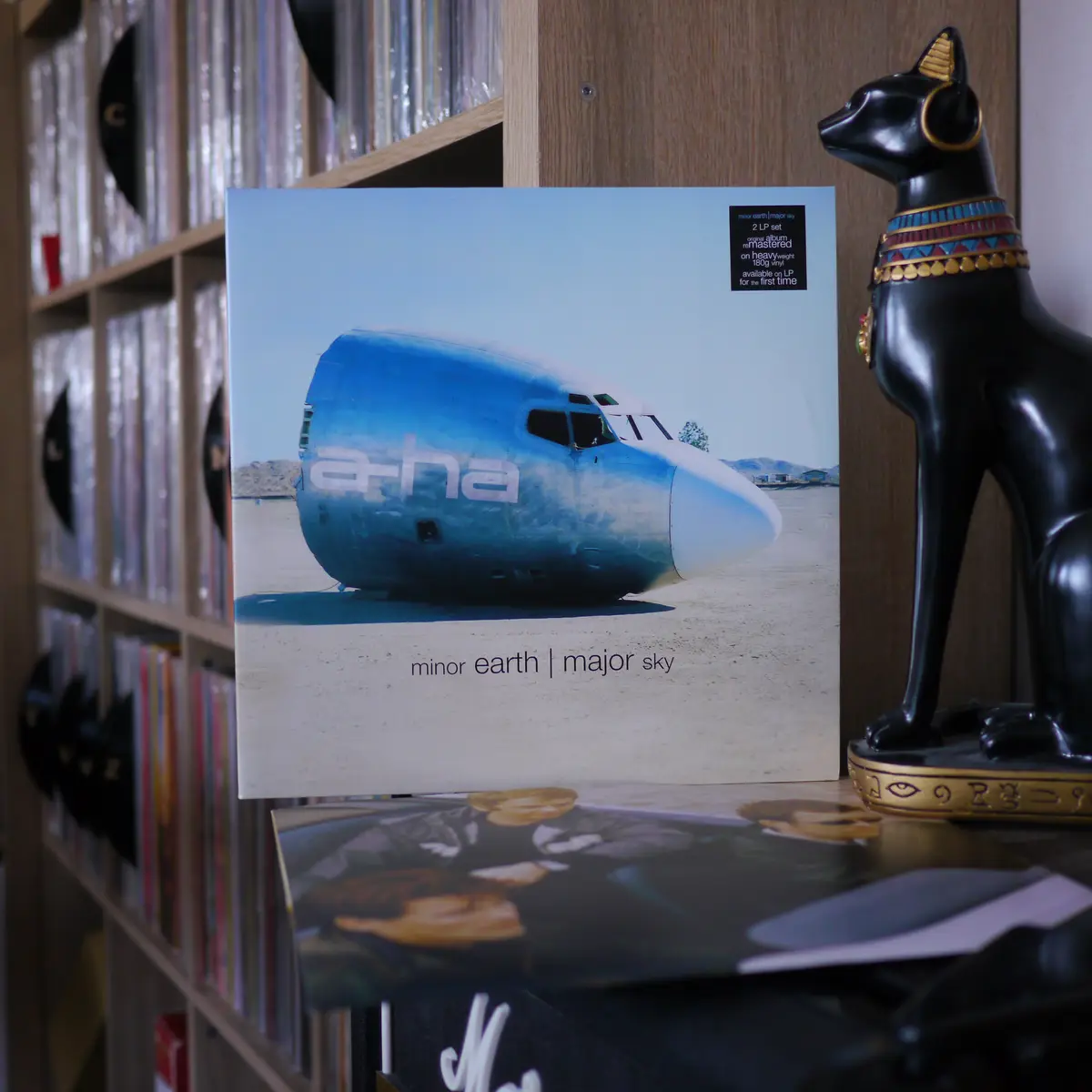A vinyl record album cover for the band a-ha's 'minor earth | major sky' is displayed on a shelf, featuring a blue airplane…