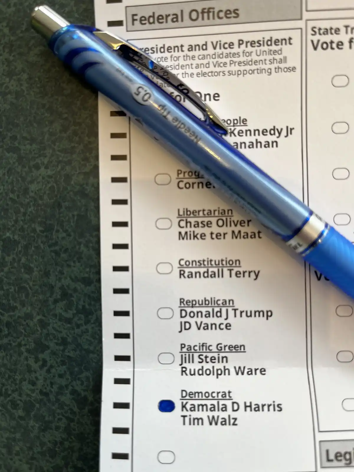 A close-up photo shows a blue pen resting diagonally across a U.S. federal ballot. The ballot lists presidential candidates…