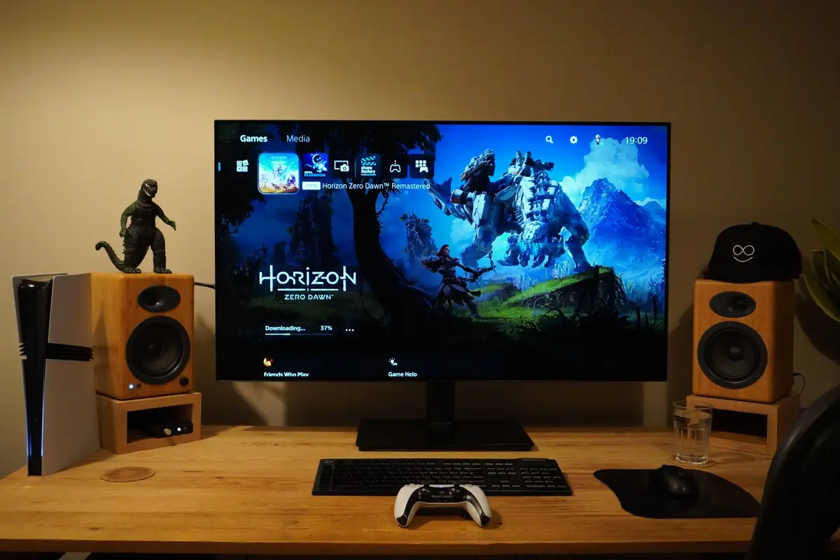 A PlayStation 5 console sits on a wooden desk next to two wooden speakers, one topped with a Godzilla figurine and the other…