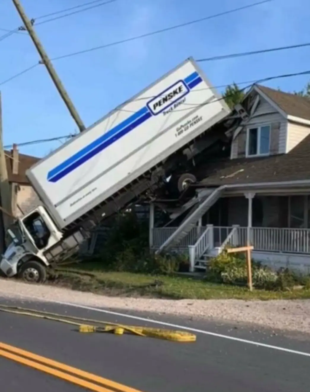 A white Penske truck rental is severely damaged, tilted at an extreme angle, with its cab on the road and trailer lodged…