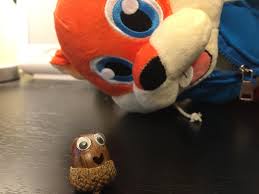 A large orange fox plushie leans over a small acorn that has been carved with googly eyes and a smiling mouth, creating a…