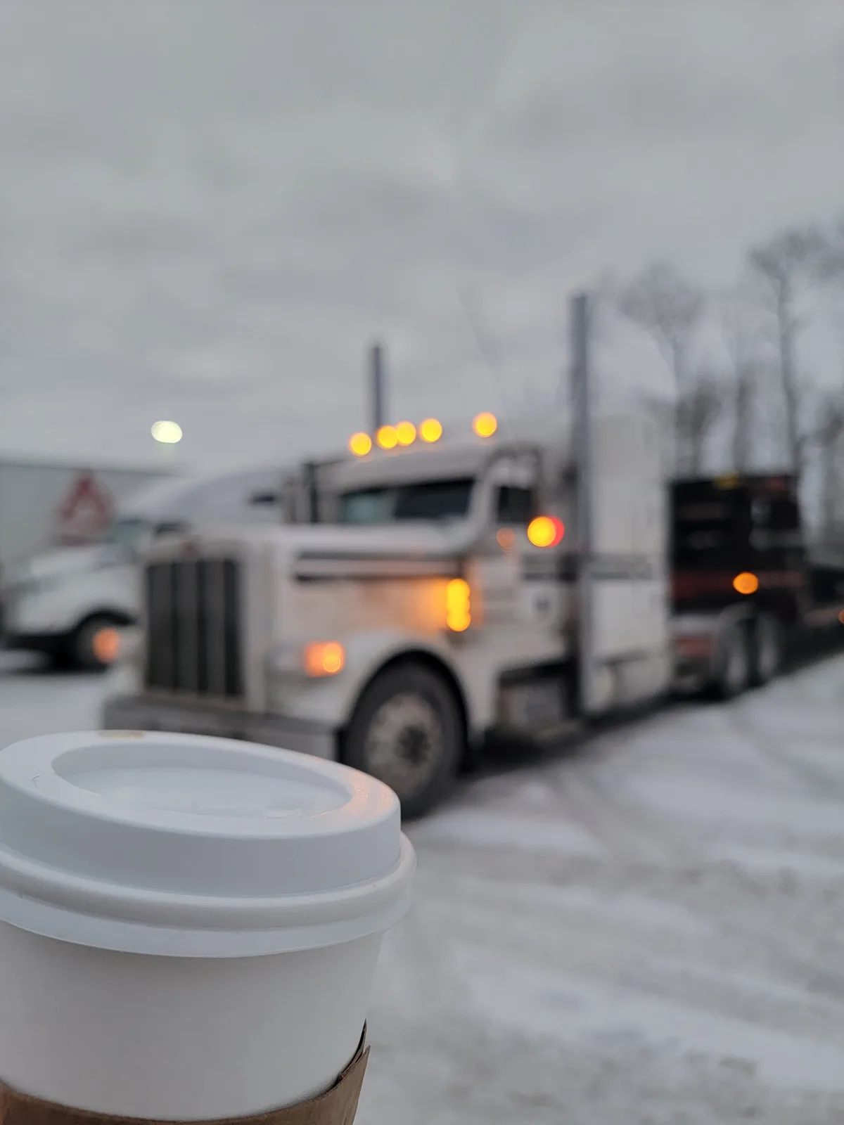 A close-up of a white disposable coffee cup with a lid is in sharp focus in the foreground, while a large white semi-truck…