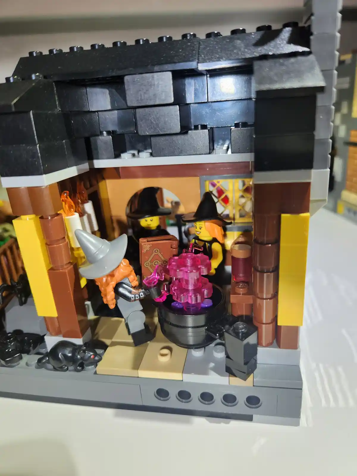 A LEGO witch's kitchen scene with three minifigures: one with a gray hat stirring a pink potion in a cauldron, another with…