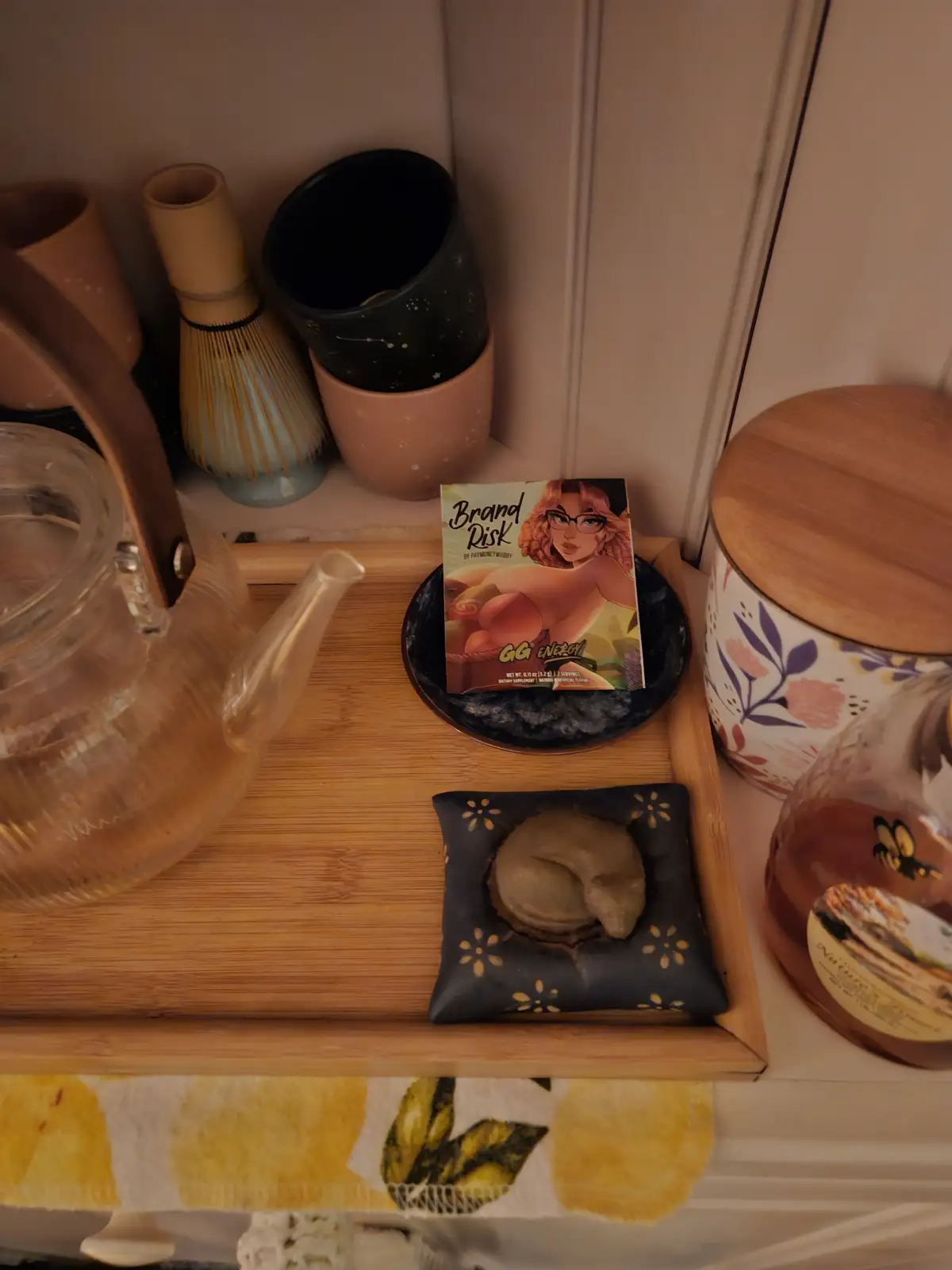 A wooden tray holds a clear glass teapot, a packet labeled 'Brand Risk GG ENERGY' with an illustration of a woman, a small…
