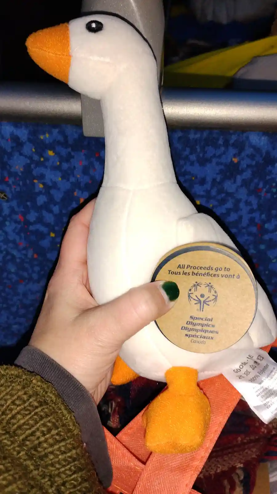 A person holds a white plush goose with an orange beak and feet, showing its circular label that reads 'All Proceeds go to…