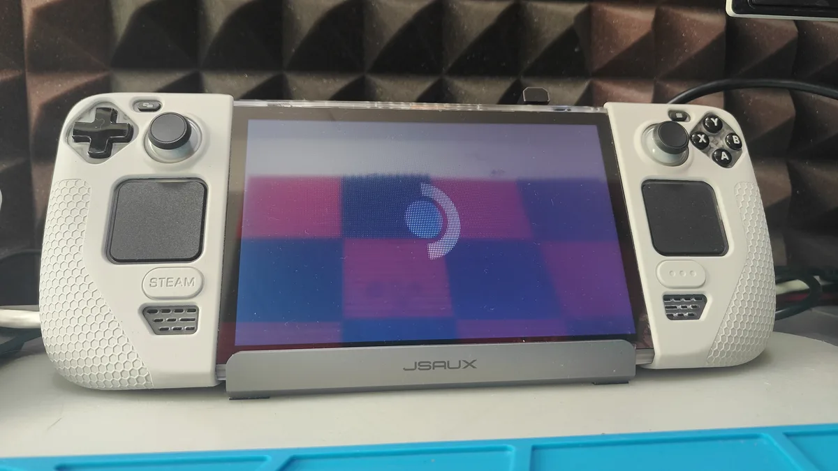 A white Steam Deck handheld gaming console sits on a stand, its screen displaying a blue loading spinner over a purple and…