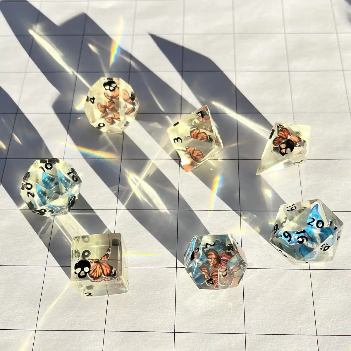 Several transparent, butterfly-embedded polyhedral dice (d20, d12, d10, d8, d6, d4) rest on a gridded surface, casting sharp…