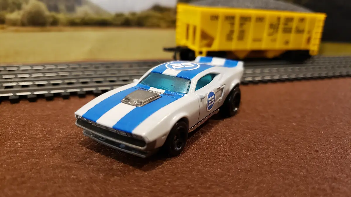 A white and blue toy race car with the number 25 on its hood and side sits on brown carpet next to model train tracks. In…