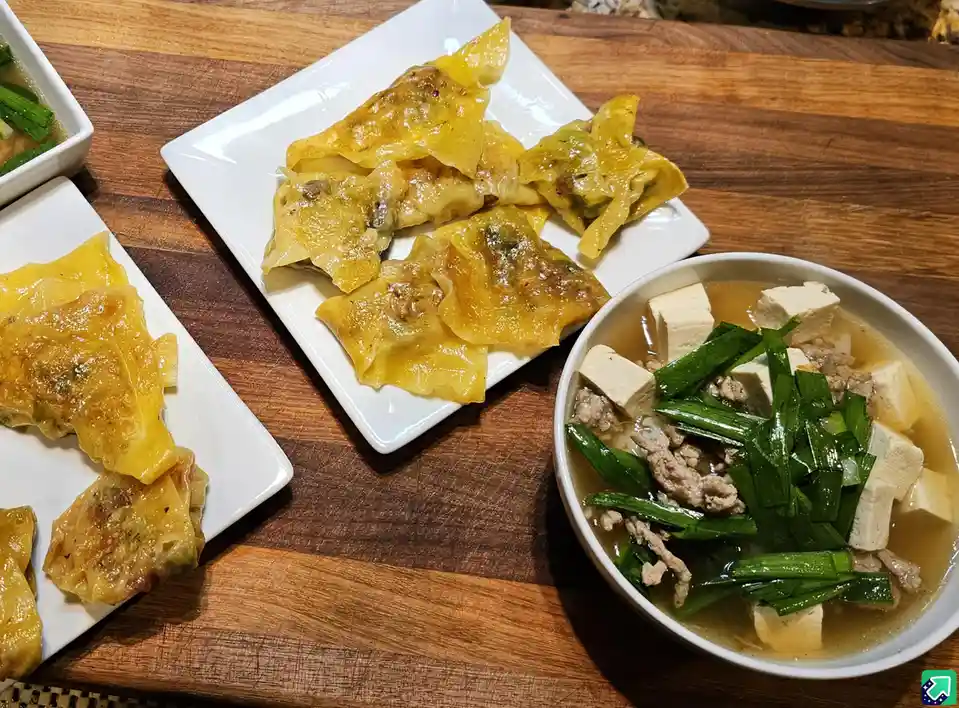 A wooden table displays three dishes: two white plates with golden-brown pan-fried dumplings and a white bowl of tofu and…