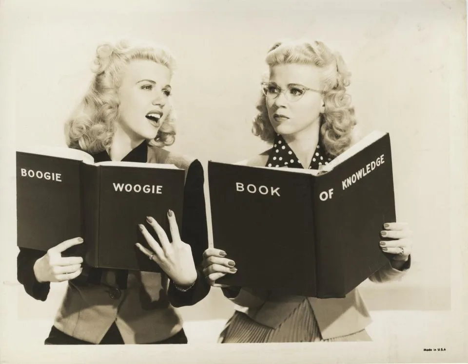 Two women in vintage 1940s-style clothing hold open books. The woman on the left, with blonde curls and no glasses, holds a…