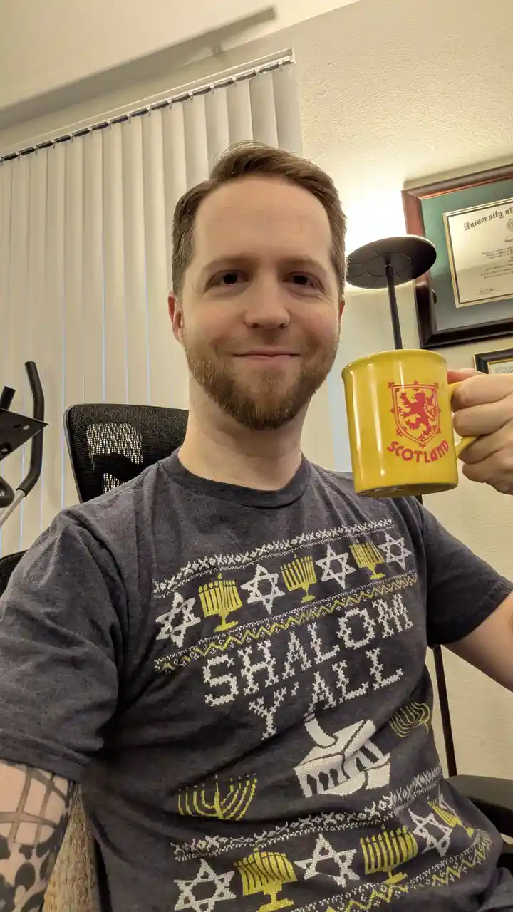 A man with a beard sits in a chair holding a yellow mug with a red Scottish lion rampant and the word 'SCOTLAND' printed on…