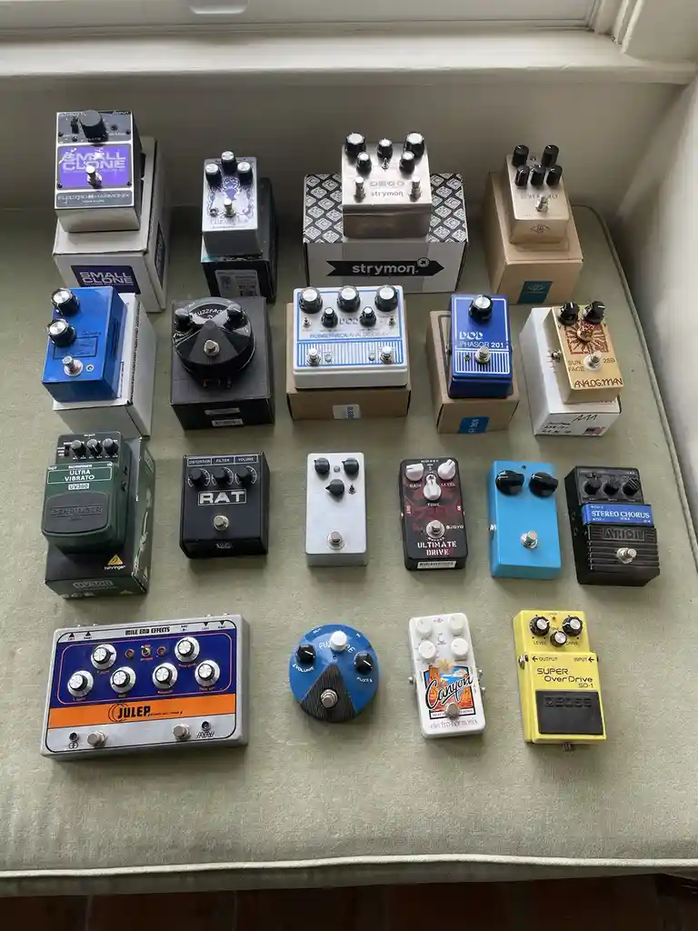 An overhead shot displays fifteen different guitar effects pedals arranged in four rows on a green fabric surface, likely a…