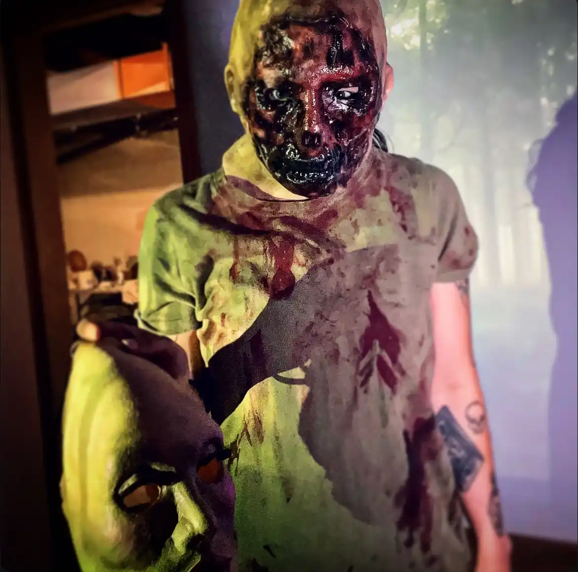 A person dressed as a zombie with a severely melted, charred, and blood-stained face, wearing a dirty green shirt also…