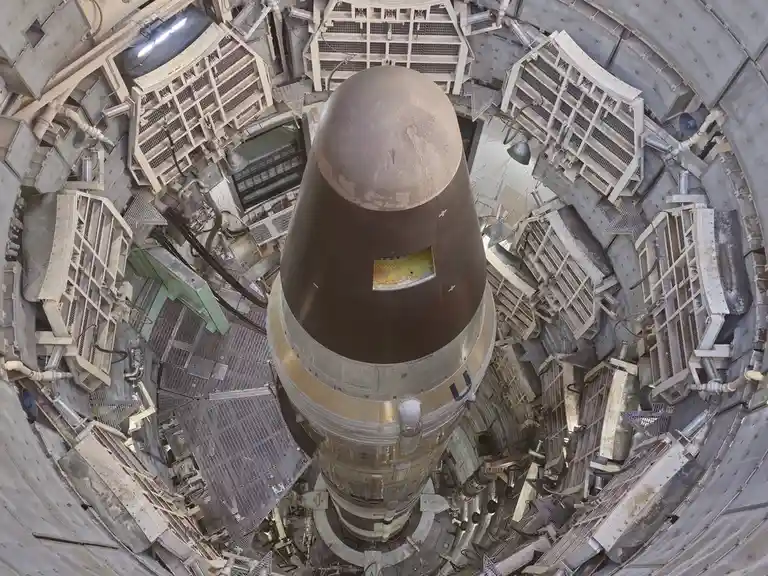 An overhead fisheye view shows a large, dark, cone-shaped missile or rocket vertically positioned in the center of a…