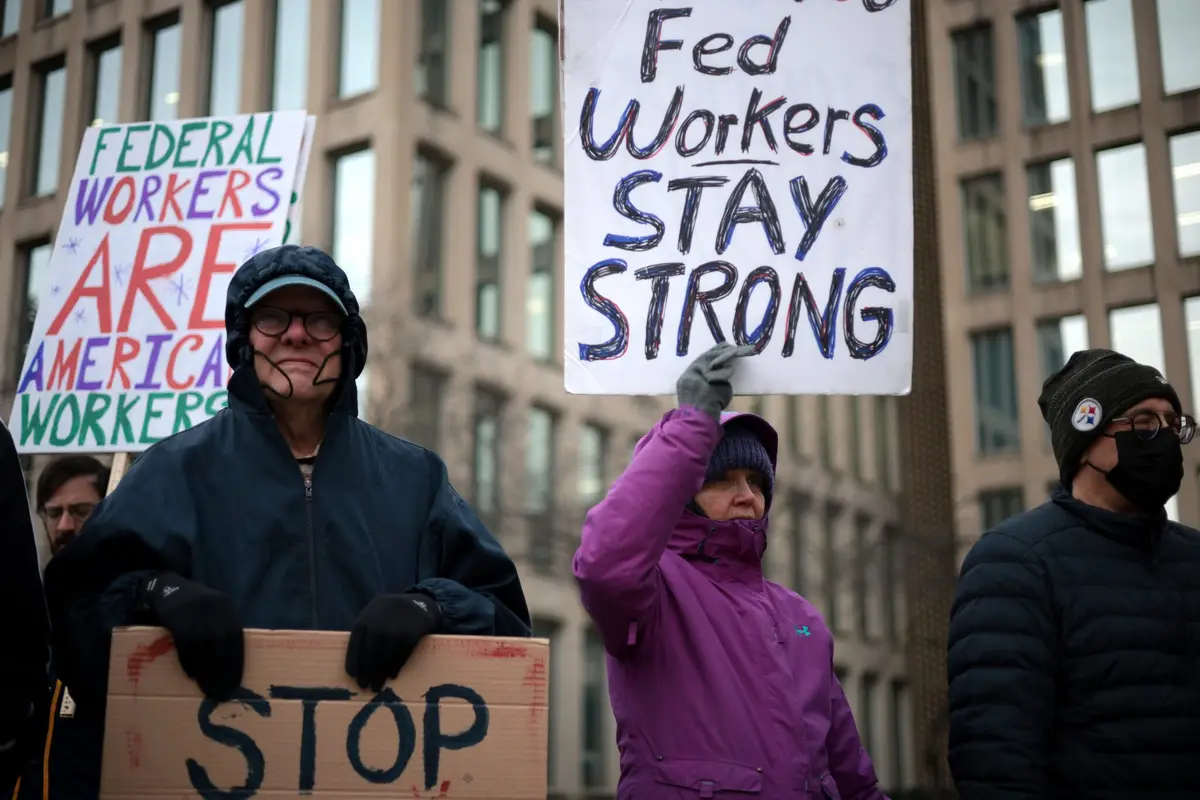 A protest scene with federal workers holding handmade signs; one reads 'Fed Workers STAY STRONG' and another 'FEDERAL…