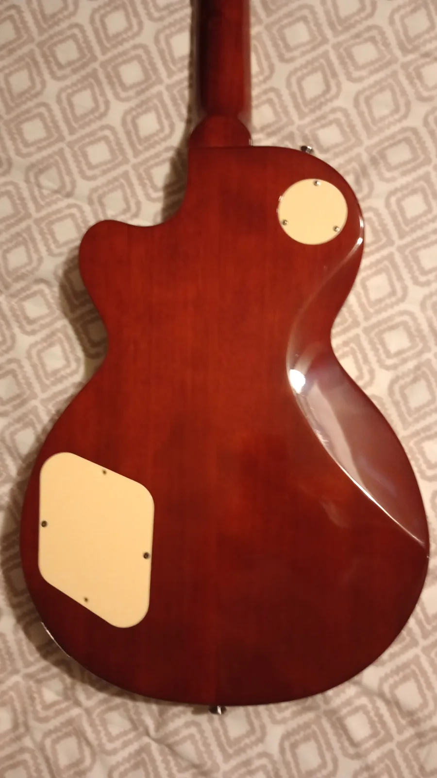 The image shows the back of a glossy, reddish-brown electric guitar resting on a patterned fabric surface. Two cream-colored…
