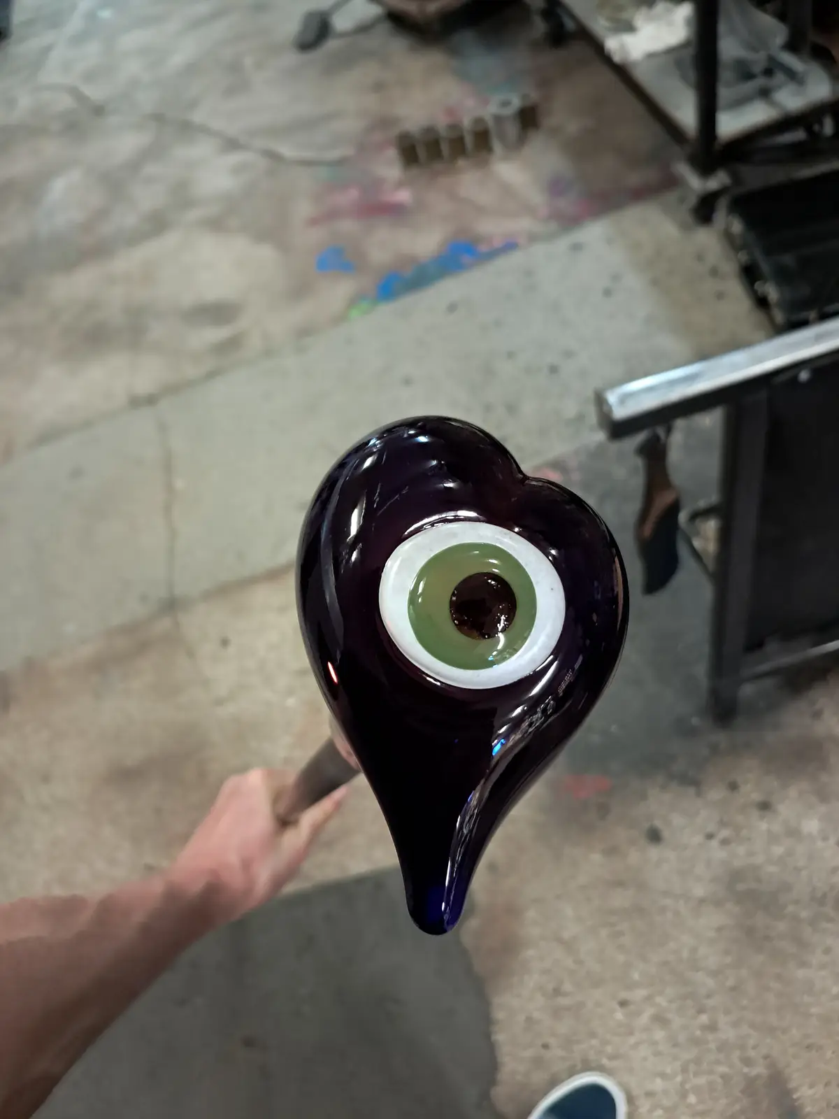 A person holds a glassblowing rod with a dark purple, heart-shaped glass piece at the end, featuring a detailed green and…
