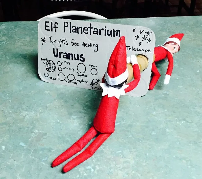 Two red-and-white elf dolls are positioned on a green countertop, one lying on its back inside a cardboard cutout labeled…