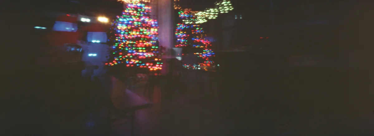 A blurry nighttime photo shows two brightly lit Christmas trees with multicolored lights, viewed through a window with…