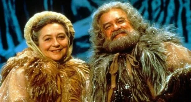 Two actors in a stage production, one woman in a golden fur-lined hood and one man with a long grey beard and fur collar…