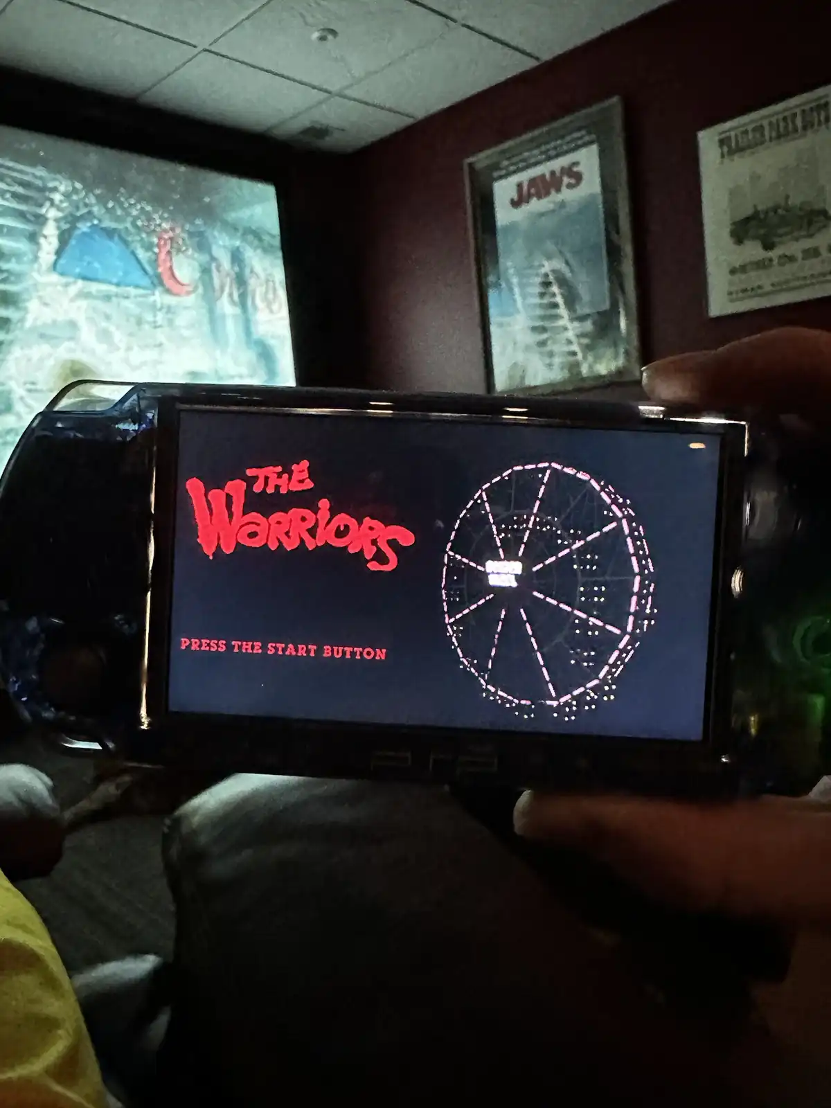 A person holds a PSP console displaying the title screen for the game 'The Warriors', which shows the game's title in red…