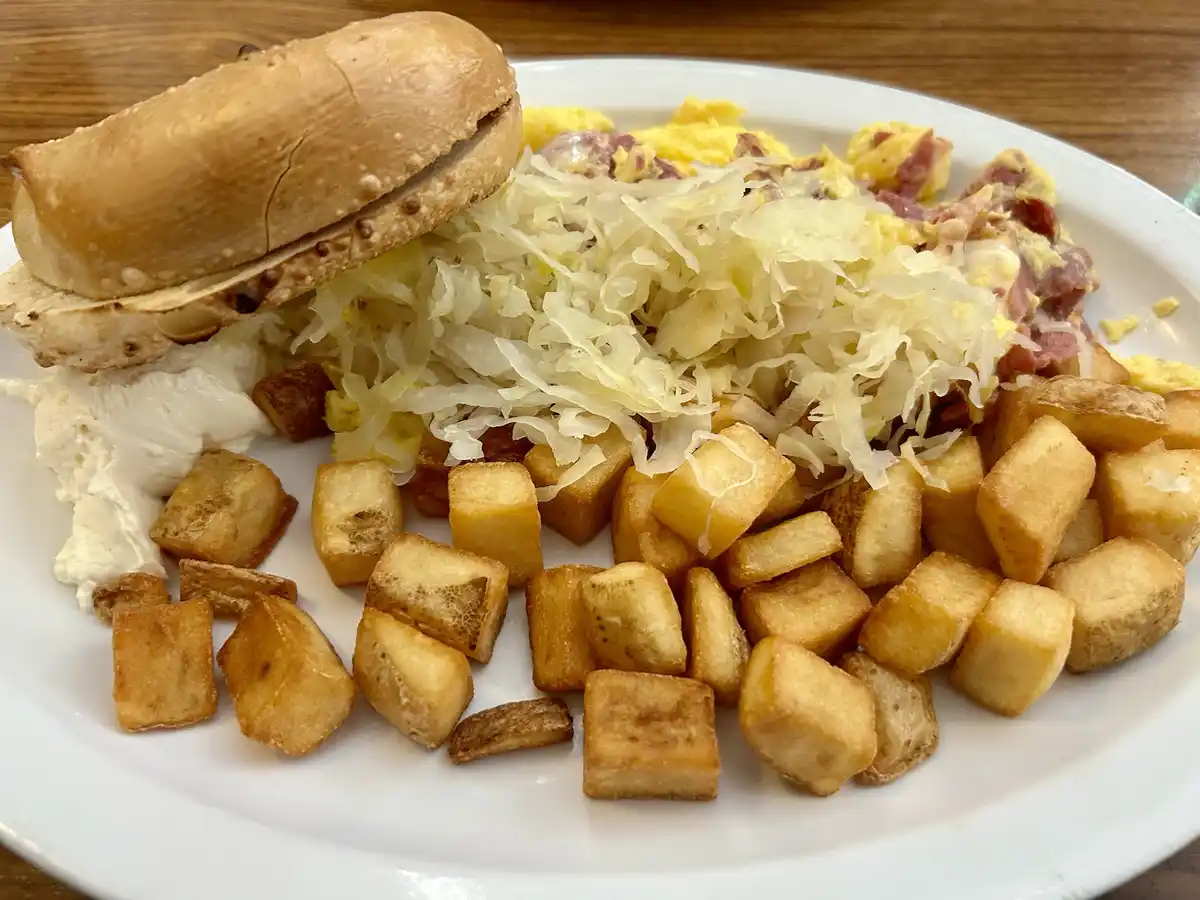 A plate of food featuring scrambled eggs with ham, a large pile of sauerkraut, crispy home fries, a half-eaten pretzel roll…
