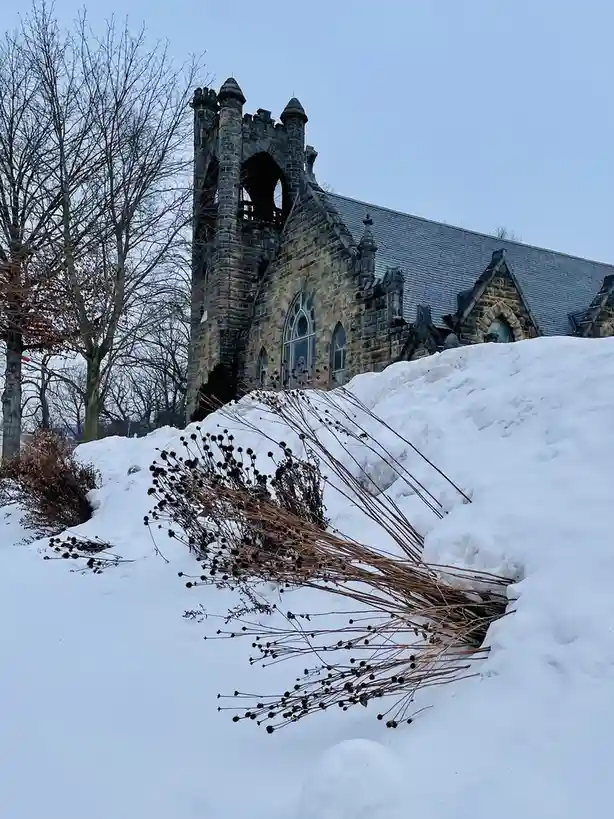 A snowy outdoor scene features a large, dark stone church with Gothic architecture, including a tower and arched windows…