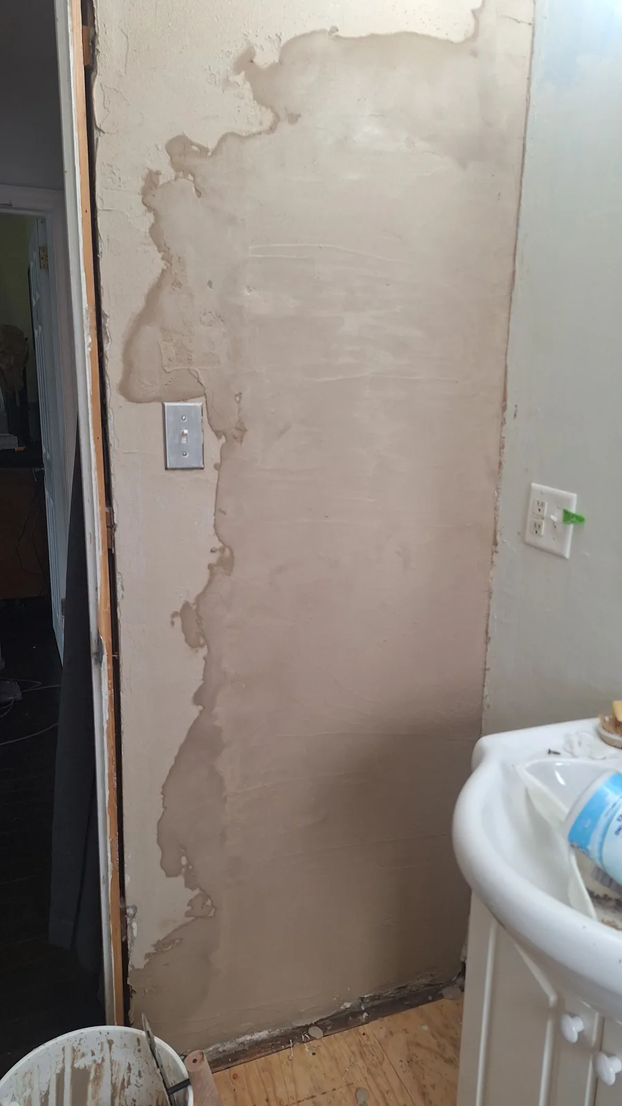A bathroom wall shows extensive damage with large patches of peeling, unevenly applied beige plaster or drywall mud…