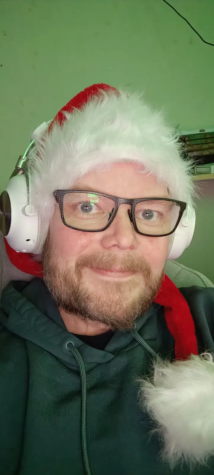 A man with a beard and glasses wears a red and white Santa hat and large white over-ear headphones while looking directly at…