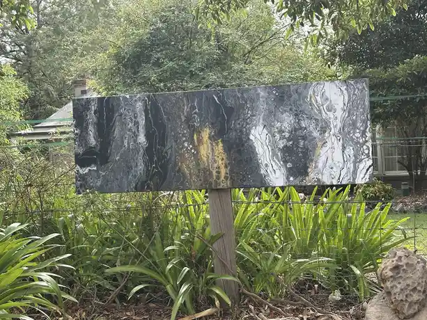A large, rectangular, abstract marbled sign with black, white, and gold swirls is mounted on a wooden post in a garden…