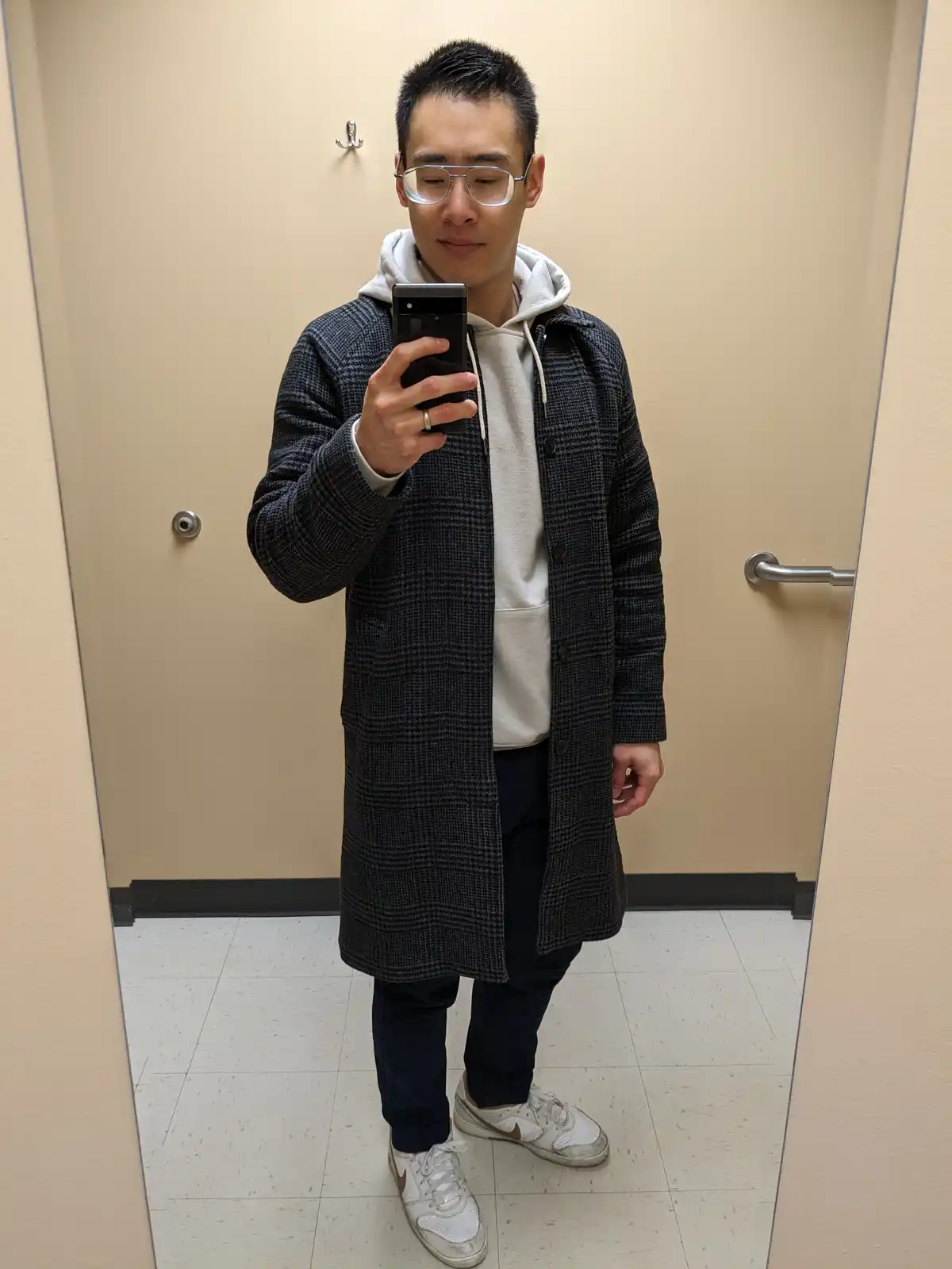 A man with glasses takes a mirror selfie in a fitting room, wearing a long dark plaid coat over a light hoodie, dark pants…