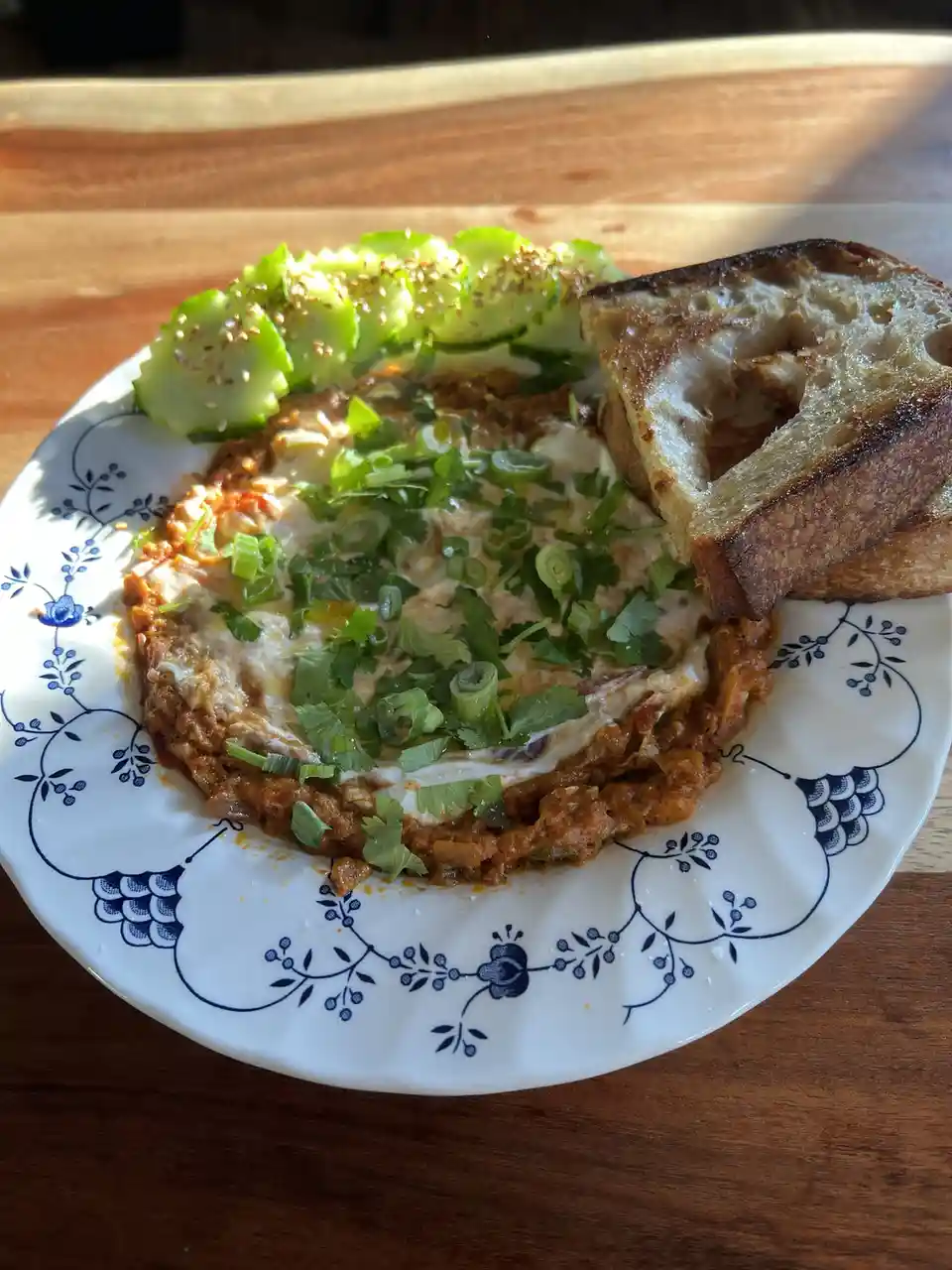 A plate of food, likely shakshuka or a similar dish, sits on a wooden table. It features a central dish with a runny egg in…