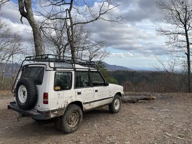 A white, muddy, older model SUV with a roof rack and spare tire is parked on a gravel overlook surrounded by bare trees. In…