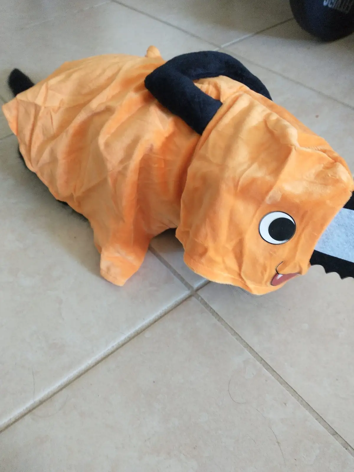 A cat wearing an orange plush costume resembling a cartoon fish with large eyes and a black fin on its back lies motionless…