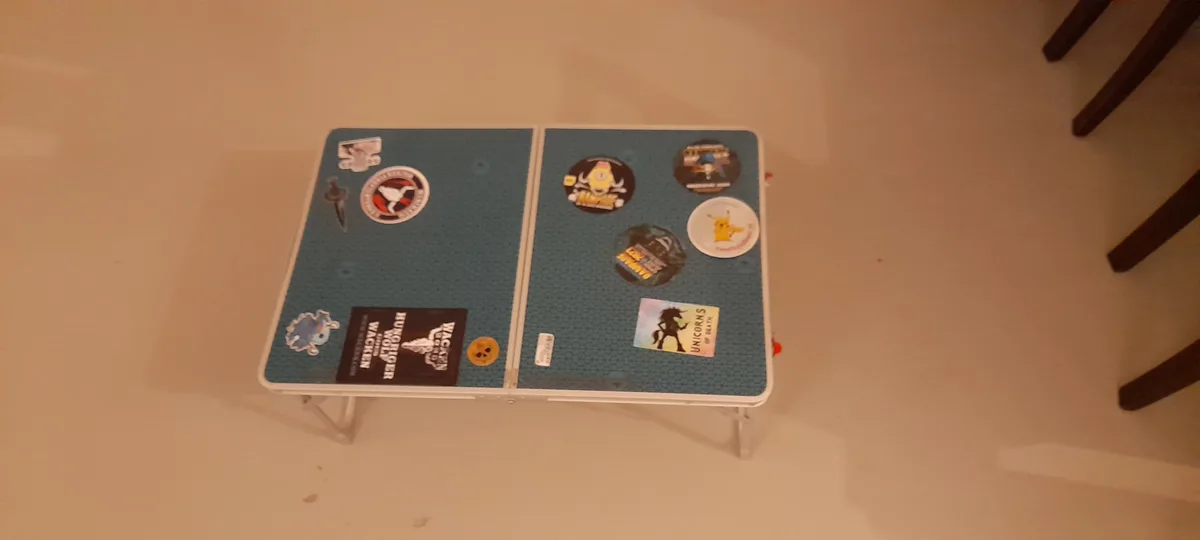 A small, foldable table with a blue textured surface is covered in various colorful stickers, including Pokémon, anime, and…