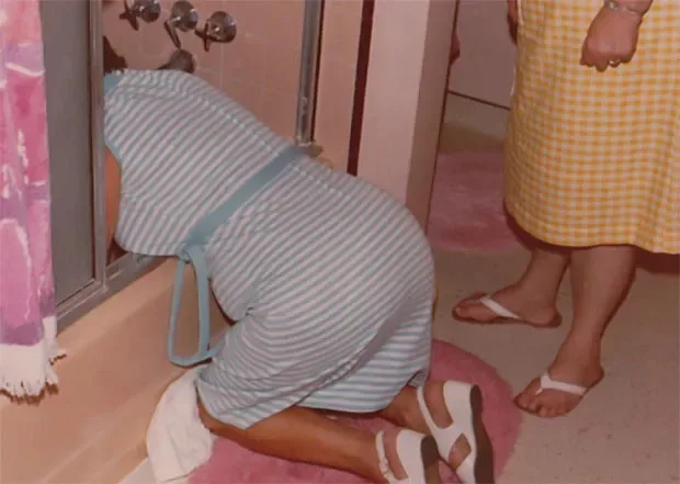 A person in a blue and white striped robe and sandals is kneeling on a pink rug in a bathroom, peering intently into a…