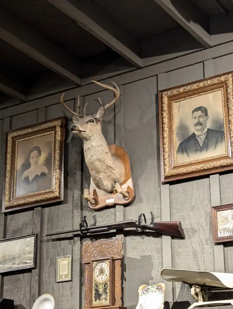 A mounted deer head taxidermy piece is centered on a wooden wall, flanked by two ornate framed portraits of a man and a…