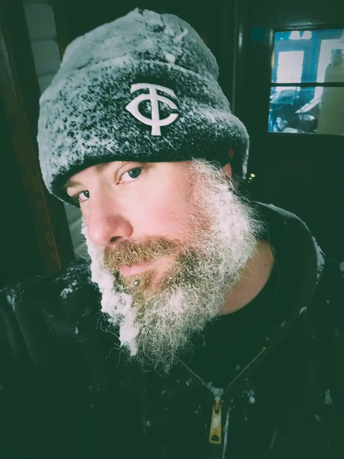 A man with a long, frost-covered beard and a snow-dusted beanie looks directly at the camera. His beard is heavily coated in…