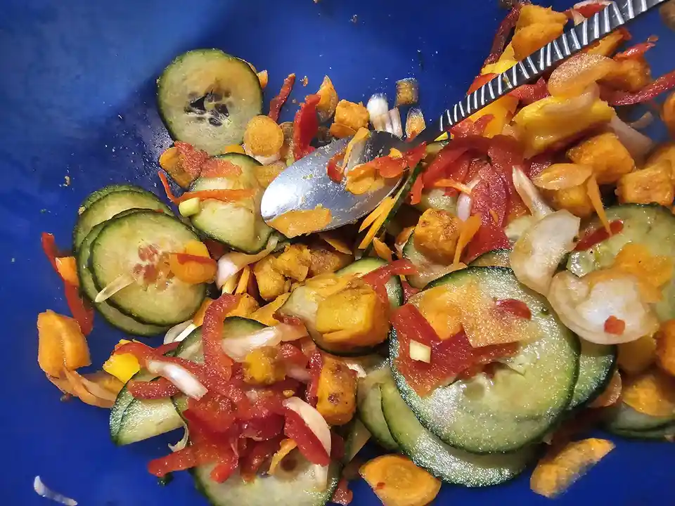 A close-up photo shows a blue bowl filled with a colorful mix of sliced cucumbers, red and yellow bell peppers, chopped…