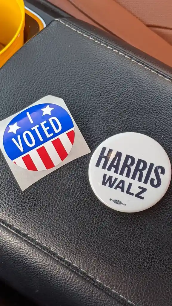 Two political items sit on a black leather car seat: a blue, red, and white 'I VOTED' sticker and a white circular button…