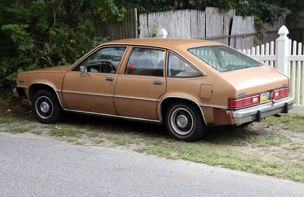 A brown, visibly aged and rusty 1970s-era sedan is parked crookedly on a grassy patch beside a paved road, with a white…
