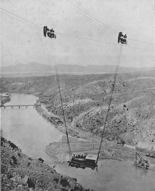 a vintage black-and-white photograph shows a cable car suspended over a wide river, with two large cable cars visible at the…