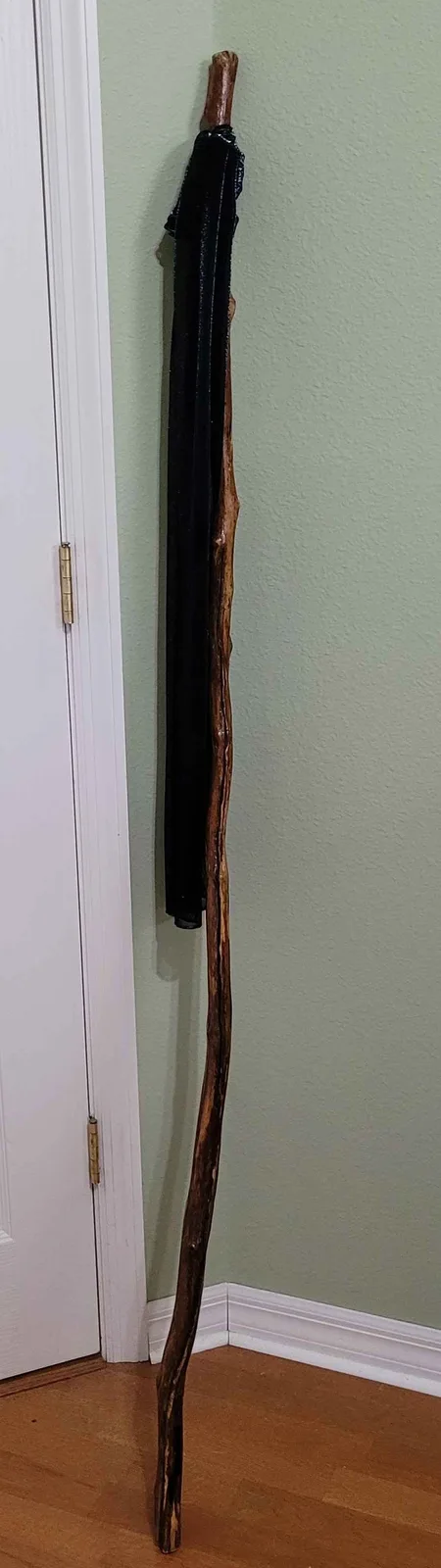 A long, irregularly shaped wooden stick leans against a light green wall next to a white door, with a black umbrella…