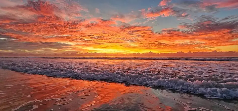 A vibrant sunset over the ocean with fiery orange, red, and yellow clouds reflecting on the wet sand and foamy waves. The…